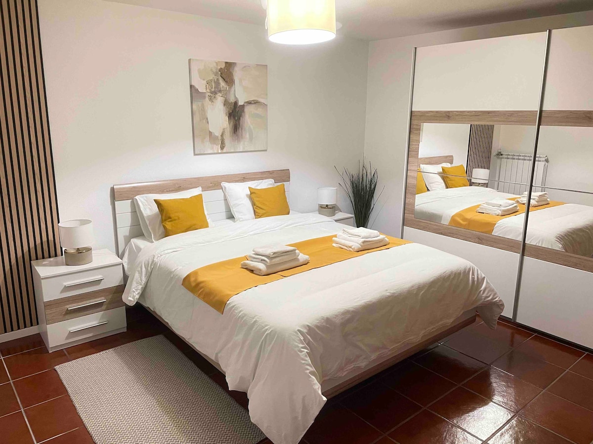 A spacious bedroom features a large bed dressed in crisp white linens with a yellow accent. Two bedside lamps illuminate the space, while a mirrored wardrobe reflects the room's design. A soft area rug lies on the tiled floor, complementing the warm color palette.