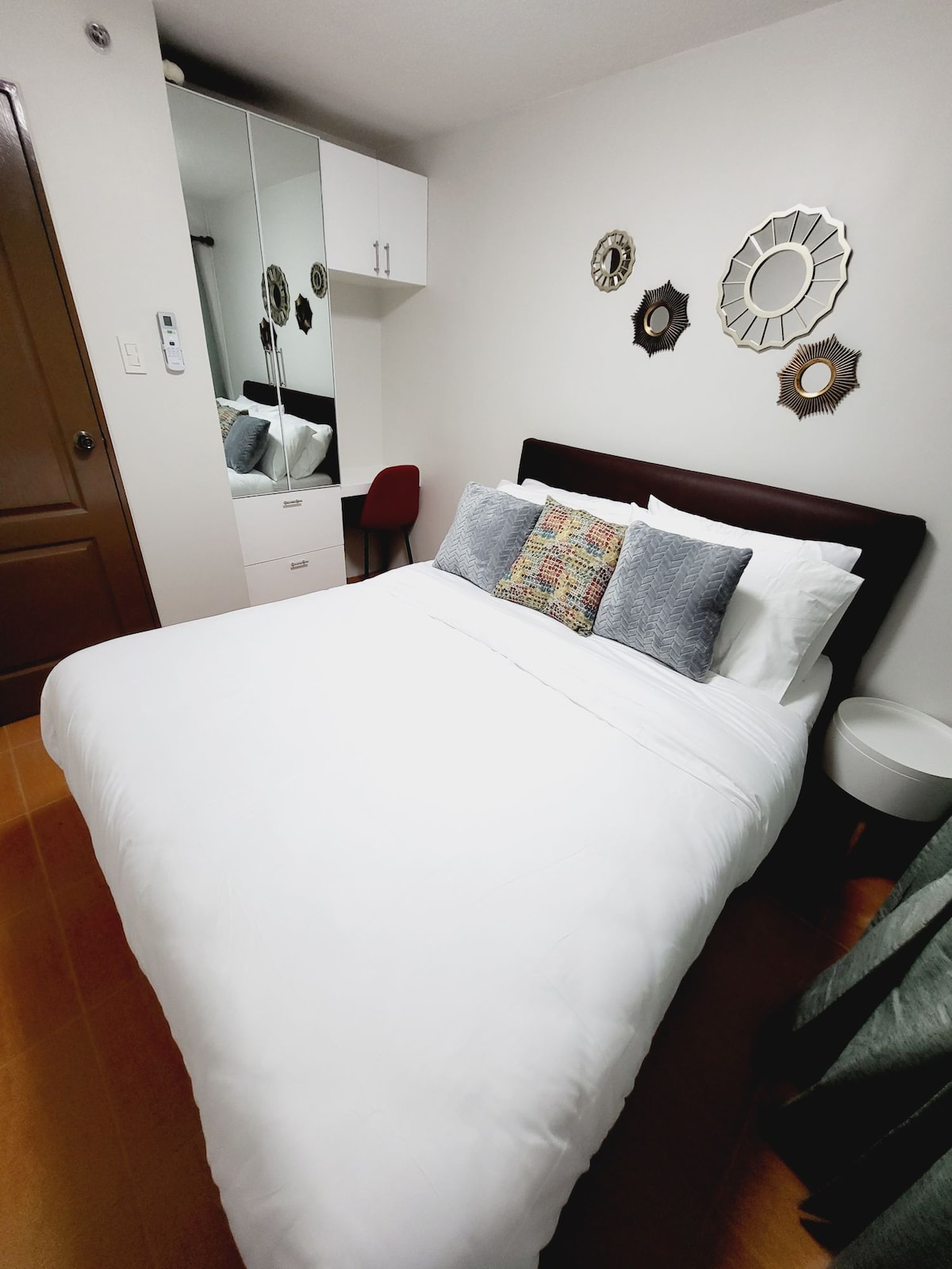A well-organized sleeping area is highlighted by a spacious bed dressed in white linens. Decorative pillows add subtle color. A mirrored cabinet provides storage, while wall-mounted ornaments enhance the room's charm. A small chair is positioned nearby, and soft lighting brightens the space.