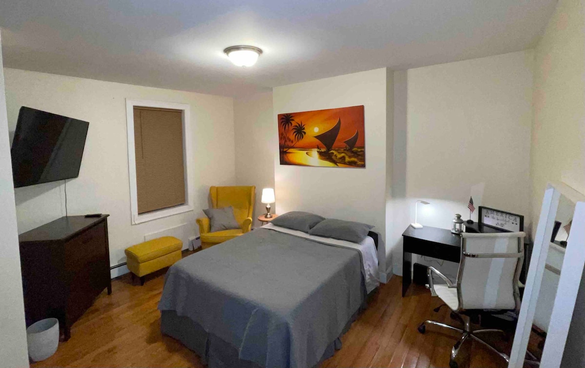 A welcoming room features a spacious bed with neutral bedding and wooden flooring. A bright yellow armchair adds a pop of color, complemented by a modern desk and chair. A television is mounted on the wall, and a serene piece of artwork decorates the opposite wall.