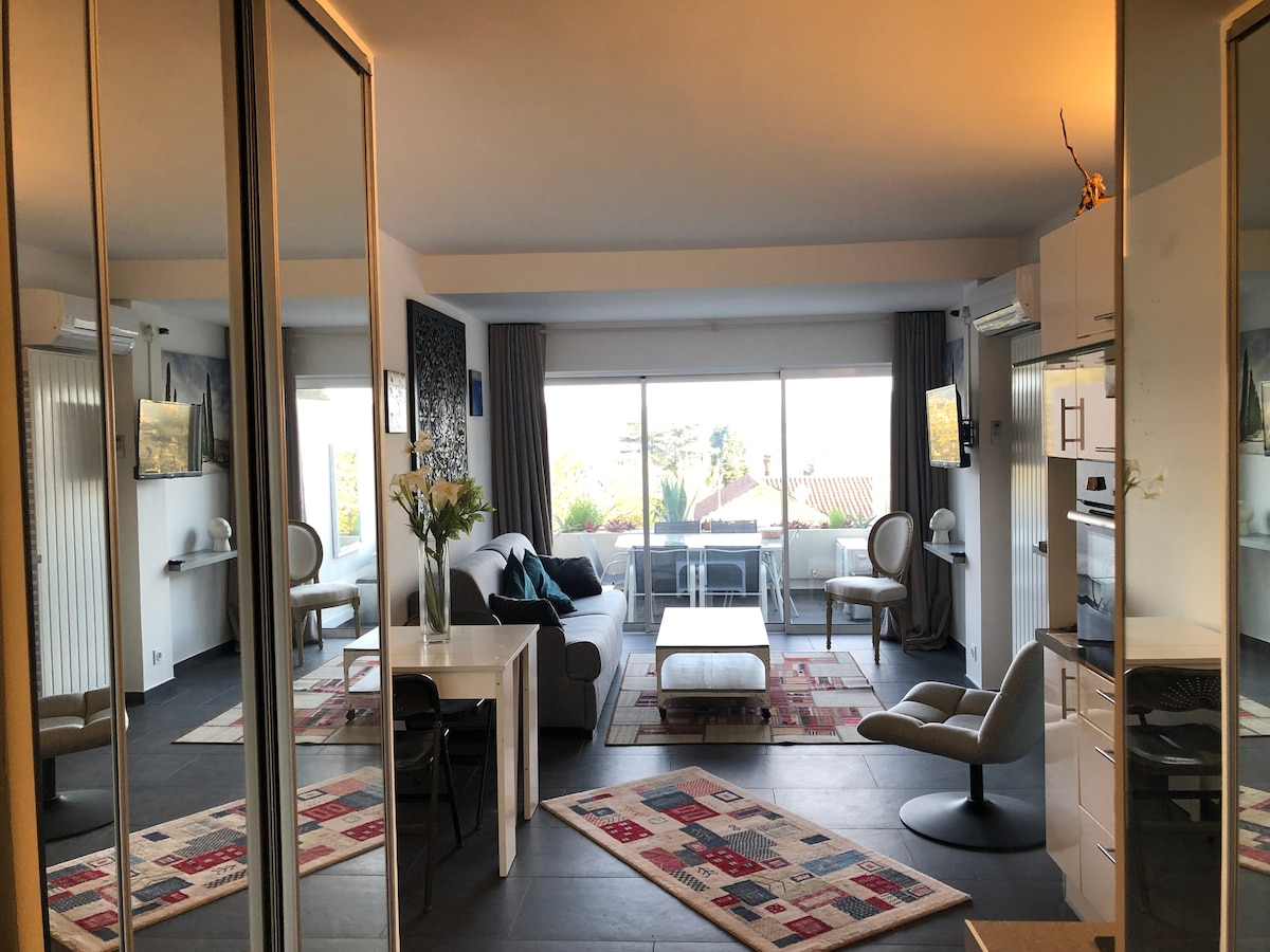 The studio features a modern living space with large windows allowing natural light to fill the room. A comfortable seating area with a light sofa is complemented by a stylish table. Mirrored walls enhance the sense of space, while decorative rugs add warmth.