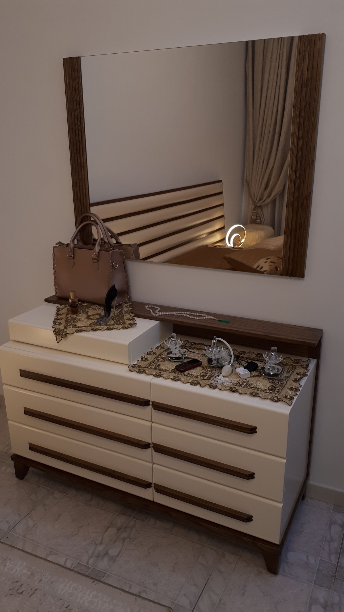 A dresser is shown topped with decorative items, including jewelry and a handbag. A large mirror reflects soft light, enhancing the contemporary design. The background features a spacious bed with a neutral headboard, and elegant curtains frame the window.
