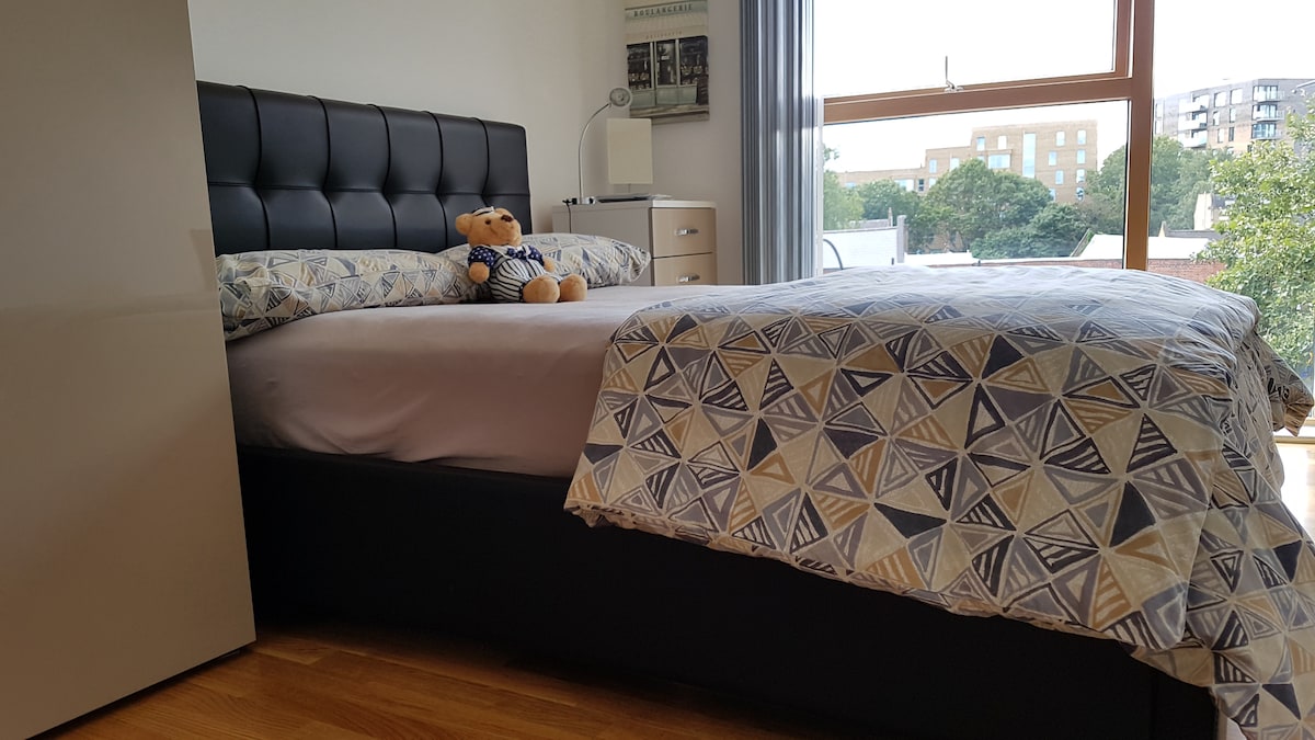 A spacious bed with an elegantly patterned duvet is positioned against a light-filled window. A teddy bear rests atop the pillows, complementing the inviting aesthetic. Nearby, a light-coloured dresser and bedside lamp are visible, providing practical storage and illumination.