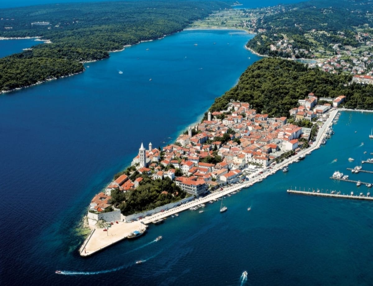 An aerial view showcases a coastal island featuring a vibrant town with terracotta roofs and a harbor filled with boats. Lush greenery surrounds the area, blending with the clear blue waters, while the shoreline reveals a mix of sandy and rocky beaches.