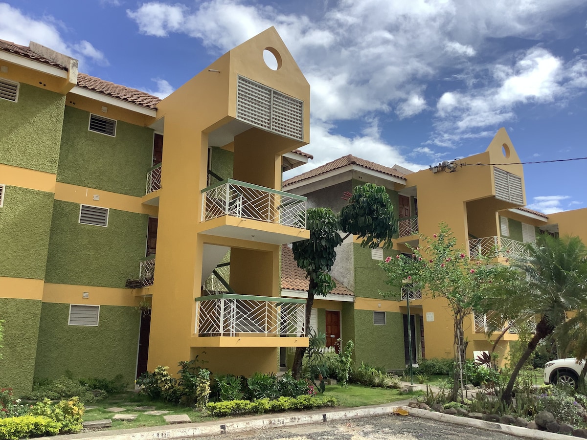 A bright, two-story building features a mix of green and yellow exteriors. Balconies are visible on each floor, framed by light railings. Lush greenery and flowering plants surround the entrance, adding a touch of nature to the inviting architecture under a partially cloudy sky.