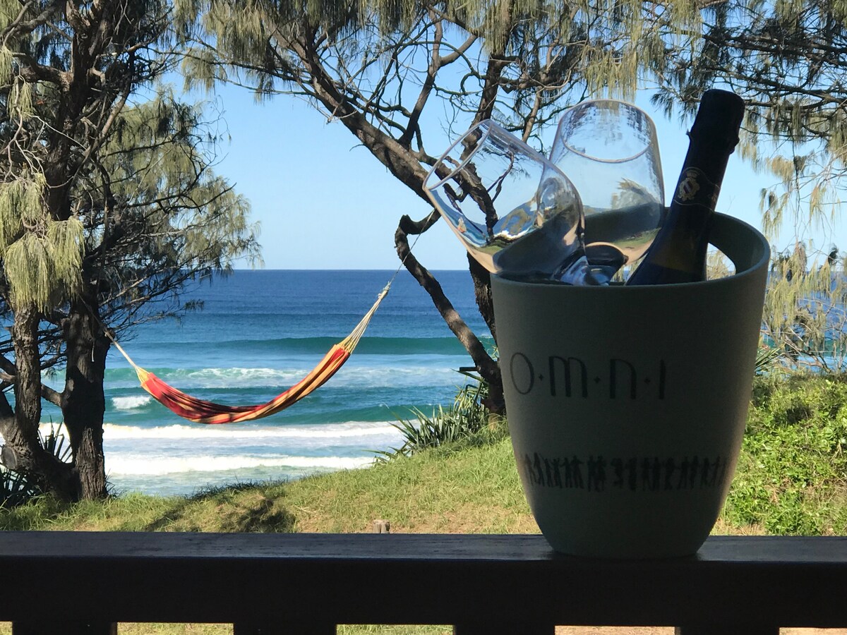 A view from the deck showcases a tranquil ocean scene, featuring rolling waves and a hammock strung between trees. In the foreground, a bucket with a bottle and two wine glasses emphasizes the relaxing atmosphere.