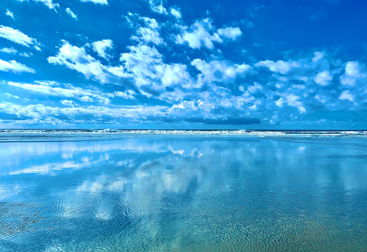 A serene ocean scene is captured, showing calm waters reflecting a vivid blue sky with scattered clouds. Gentle waves can be seen approaching the shoreline, creating a tranquil atmosphere that highlights the expansive beauty of the beach.