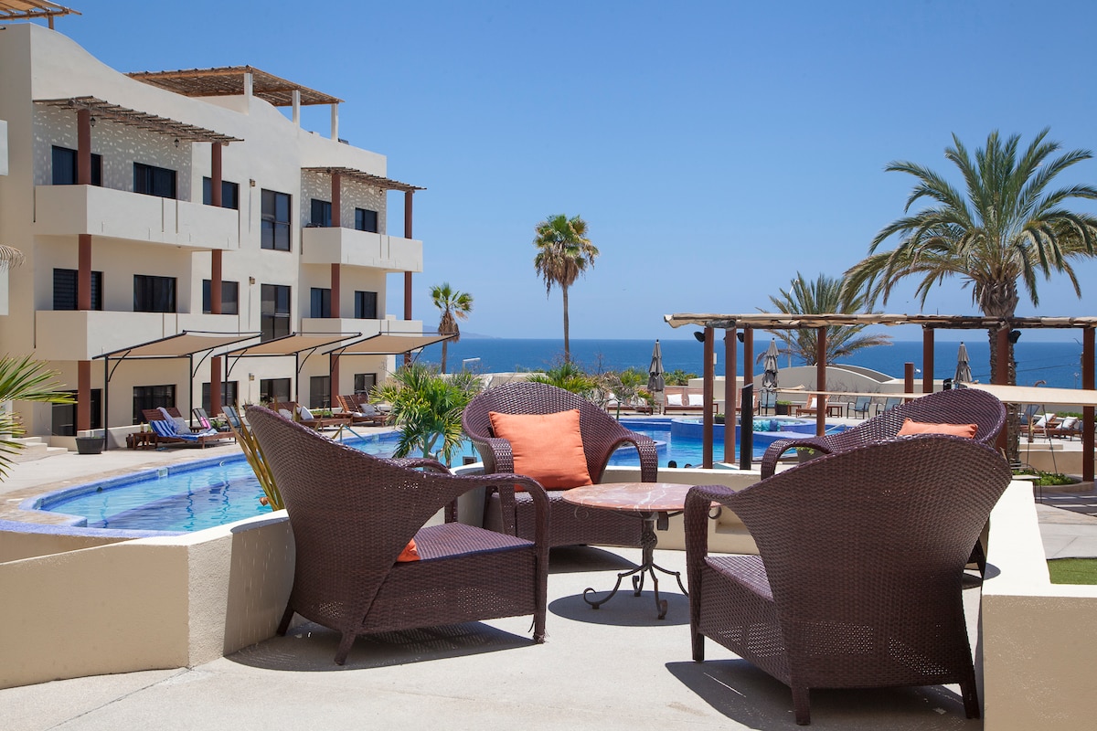 Outdoor lounge chairs are arranged around a small table, offering a view of the swimming pool and ocean in the background. Palm trees and sun umbrellas provide a tropical feel, while the three-story building features balconies and large windows overlooking the pool area.