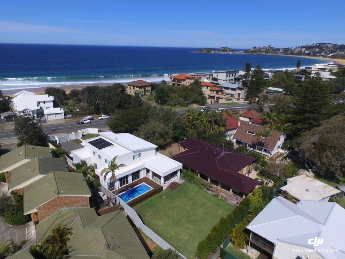 An aerial view captures the proximity of Catalina to Wamberal Beach, showcasing the home’s spacious lawn and in-ground pool. Nearby houses are visible, along with the coastline, highlighting the beach lifestyle and natural beauty of the surrounding area.