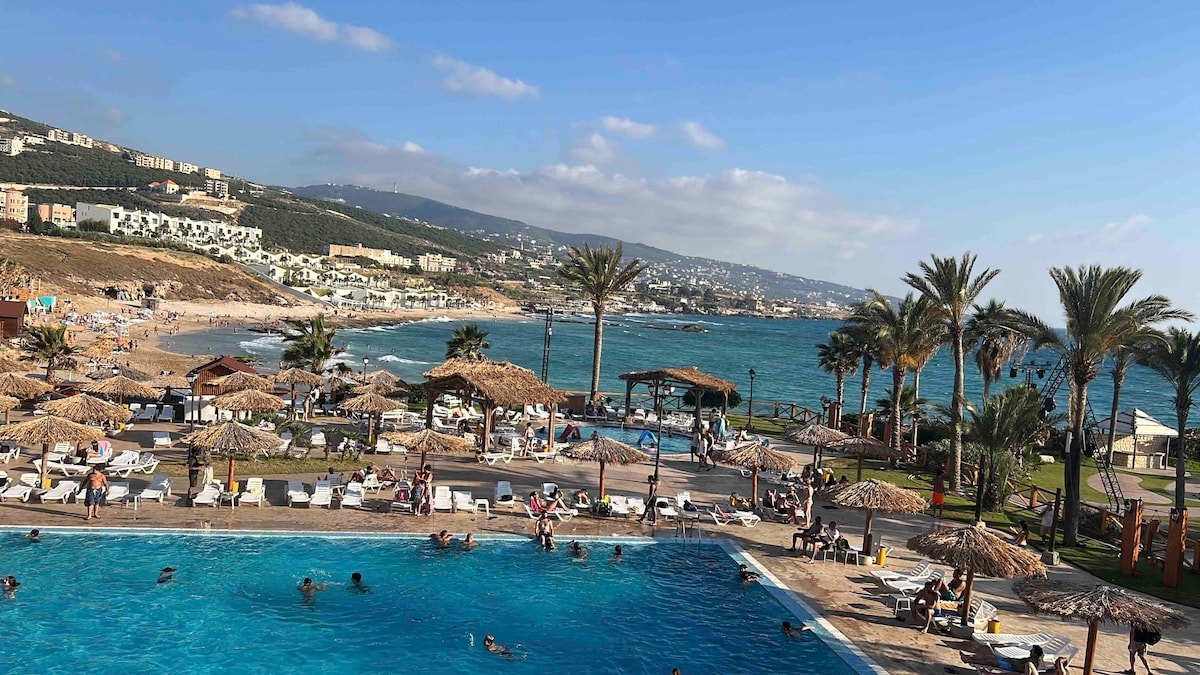 A large swimming pool is visible in the foreground, surrounded by sun loungers. Beyond the pool, a sandy beach stretches along the coastline, with people enjoying the sun. Palms sway gently near thatched-roof huts, and rolling hills are seen in the background.