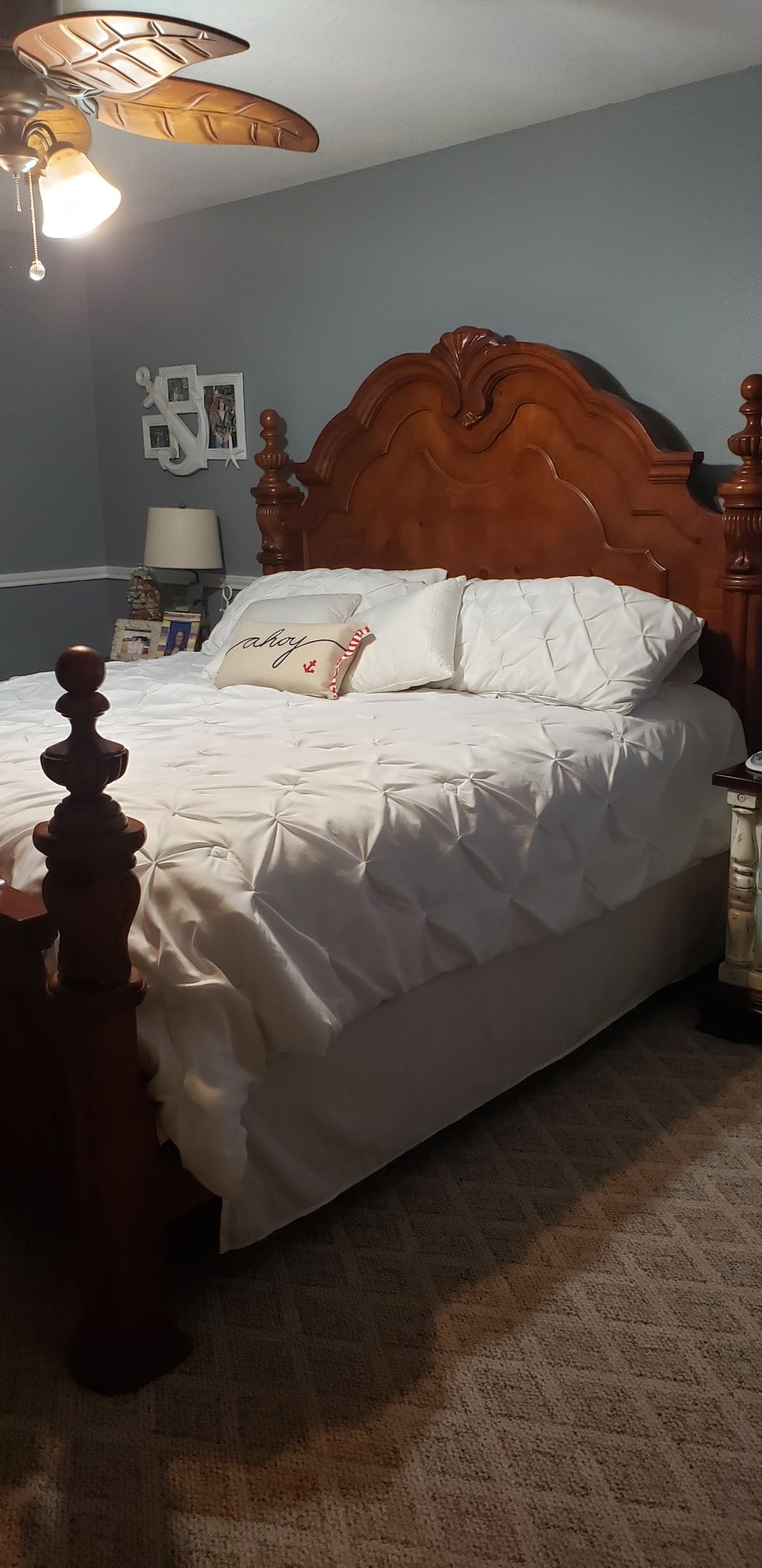 A well-furnished bedroom features a carved wooden bed frame with a plush white comforter and decorative pillows. Warm lighting is provided by a ceiling fan and bedside lamp. The space is complemented by a neutral-colored wall and carpet, creating a calming environment.