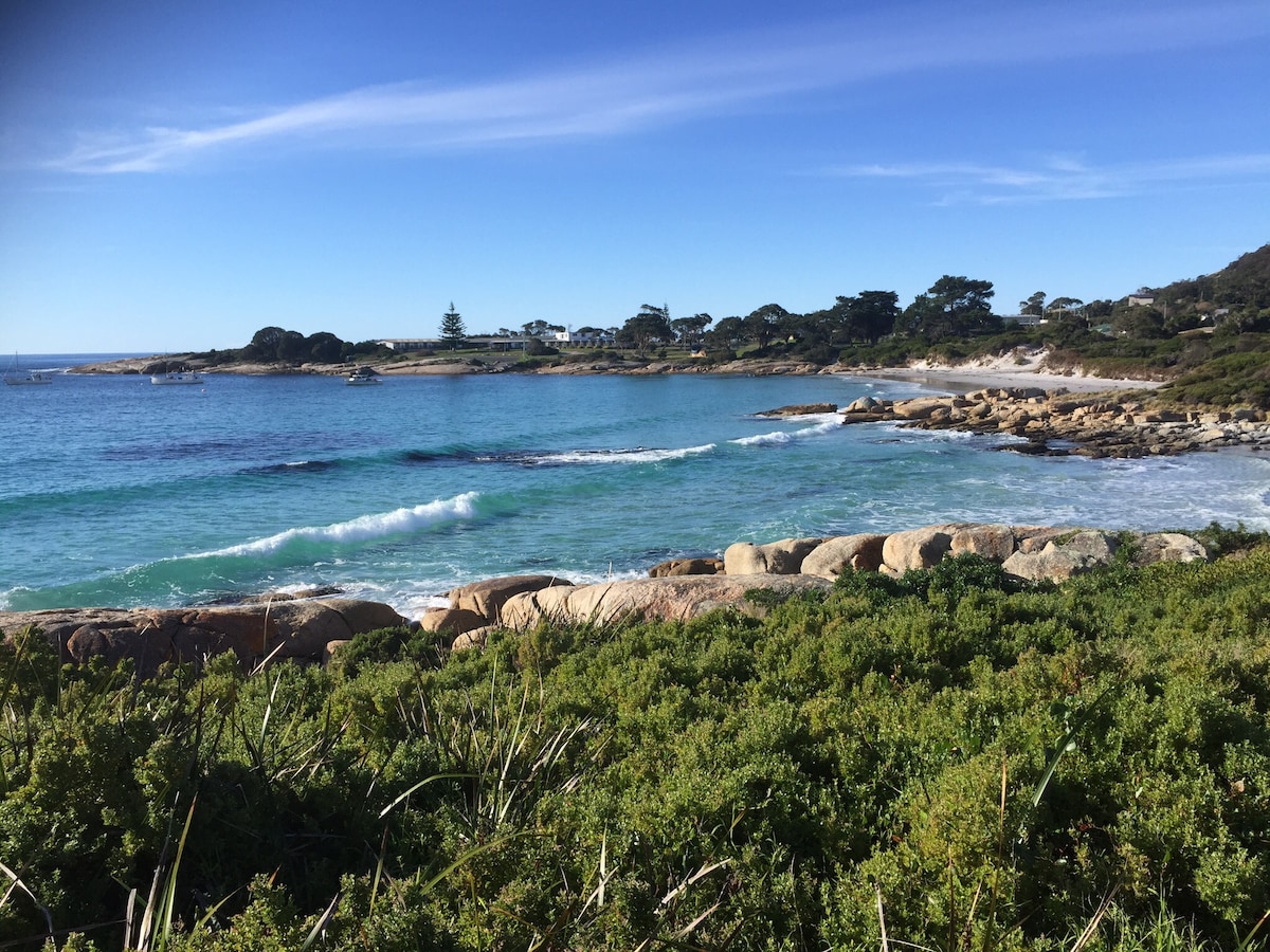 Coastal scenery features clear turquoise waters gently lapping against rocky shores. Lush green shrubs line the foreground, while the horizon displays gentle waves and distant coastal vegetation. The sky is a brilliant blue with wispy clouds, enhancing the tranquility of the beach setting.