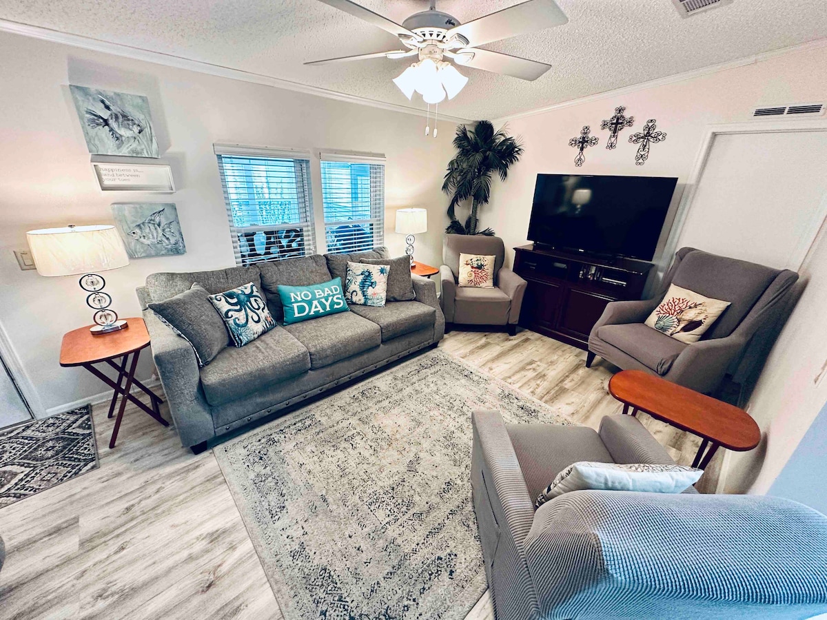 A cozy living room features comfortable seating with throw pillows, a ceiling fan overhead, and a mounted television. Natural light filters through multiple windows, while a decorative area rug complements the light wood flooring.