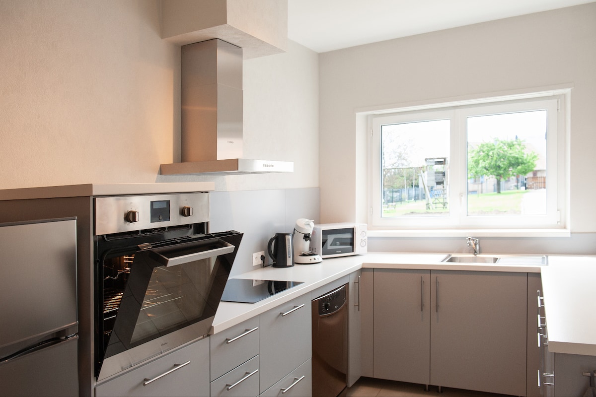 A modern kitchen is equipped with stainless steel appliances, including an oven, microwave, and refrigerator. Countertops are finished in a light color, and a window provides natural light and a view of the greenery outside.
