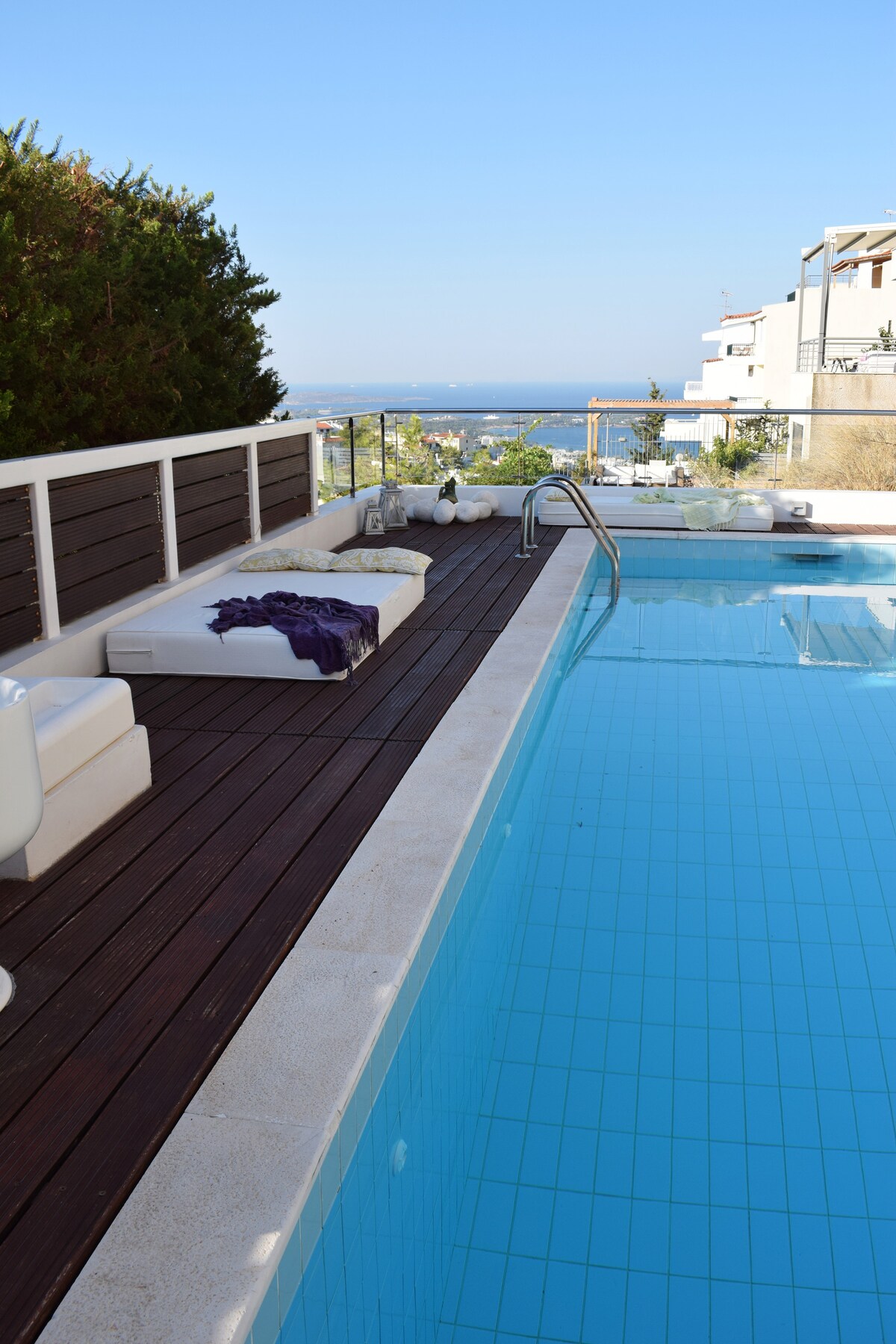 A serene swimming pool extends alongside a wooden deck, offering lounge seating with soft cushions. The clear blue water reflects the sky, while a gentle sea breeze is suggested by the surrounding greenery and distant ocean view.