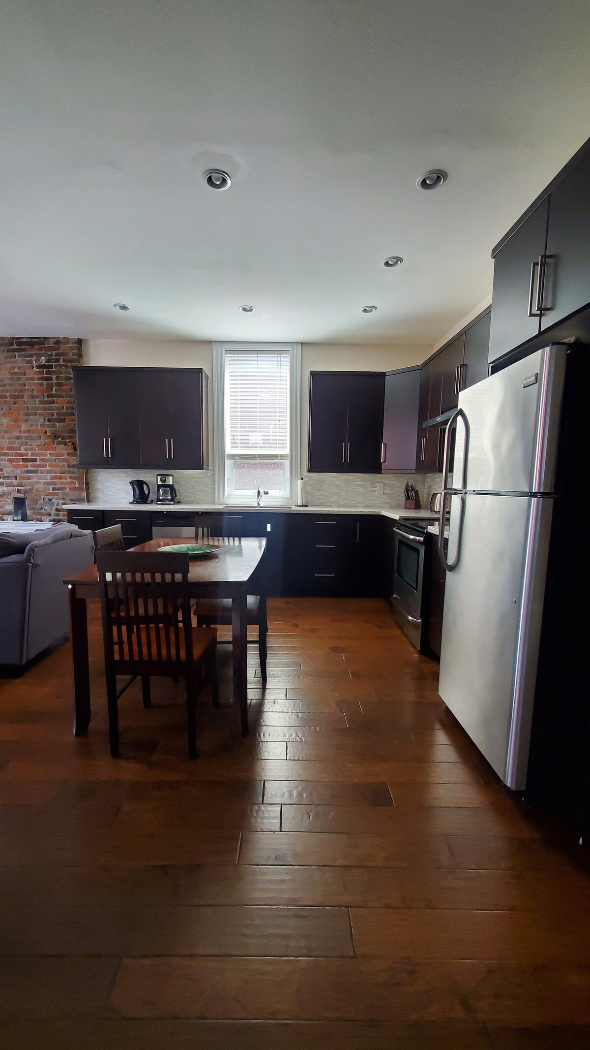 The kitchen showcases dark cabinetry complemented by a light stone backsplash. A wooden dining table with four chairs is positioned centrally. Stainless steel appliances, including a refrigerator and stove, are visible, along with a window allowing natural light to fill the space.