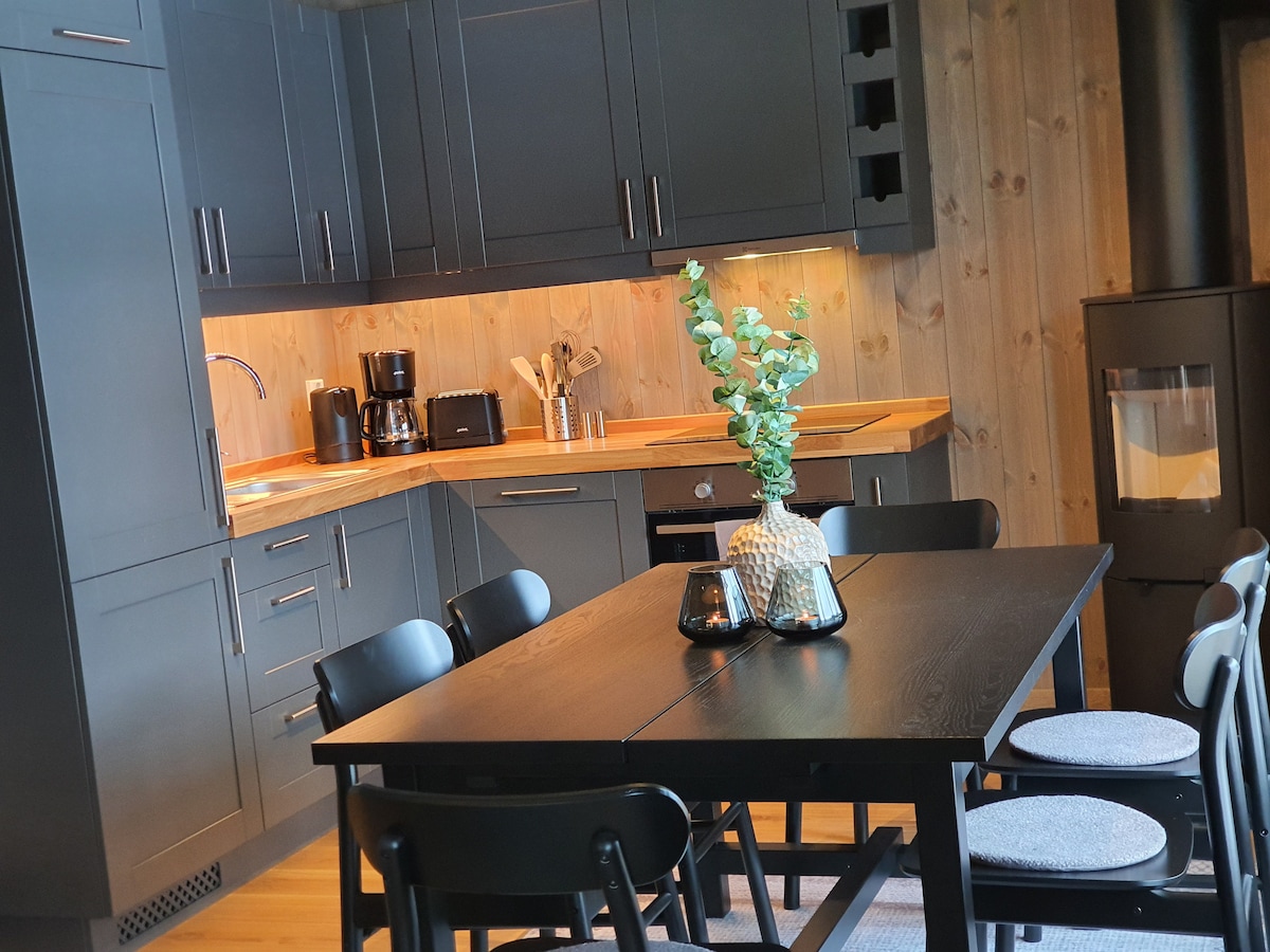 A modern kitchen is shown with dark cabinetry and wooden accents. A dining table with black chairs is set for meals, while kitchen appliances, including a coffee maker, are arranged on the countertop. A decorative plant centerpiece enhances the inviting design.