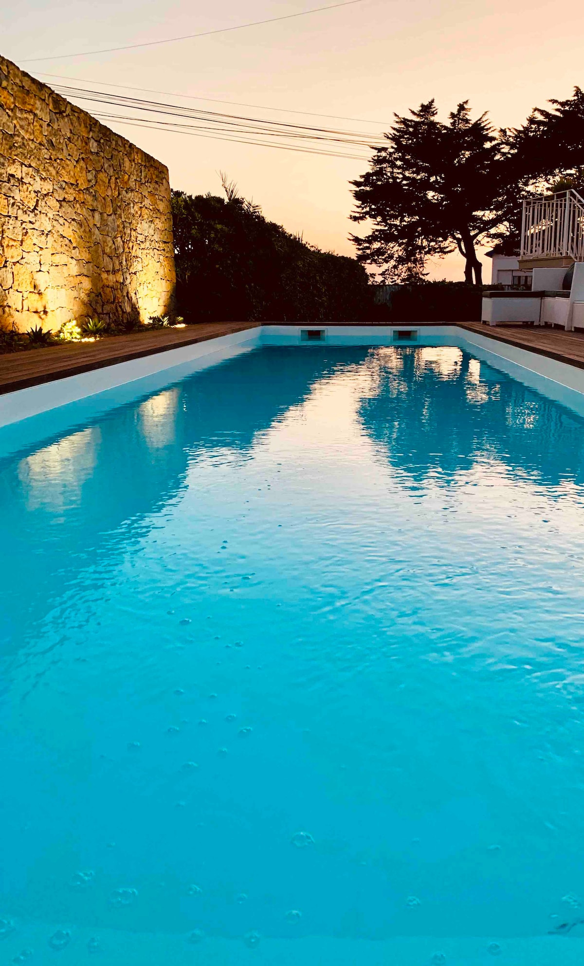 The heated pool is surrounded by natural stone walls, reflecting the soft evening light. The clear, turquoise water is calm, bordered by wooden deck areas, and illuminated by subtle uplighting along the stone, creating a peaceful atmosphere for relaxation.
