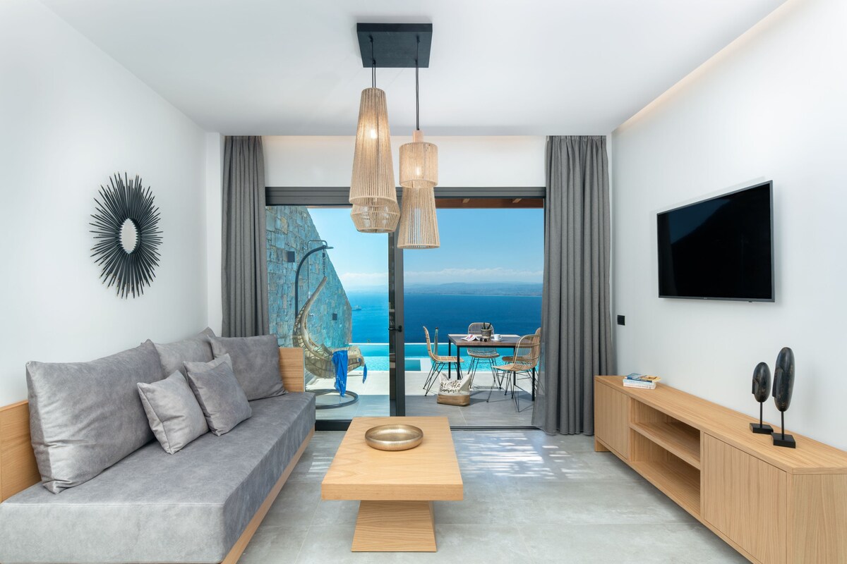The open-plan living area features a comfortable corner sofa adorned with pillows, complemented by a wooden coffee table. A 50'' HDTV is mounted on the wall. Floor-to-ceiling glass doors provide stunning sea views and access to the dining terrace, illuminated by hanging light fixtures.