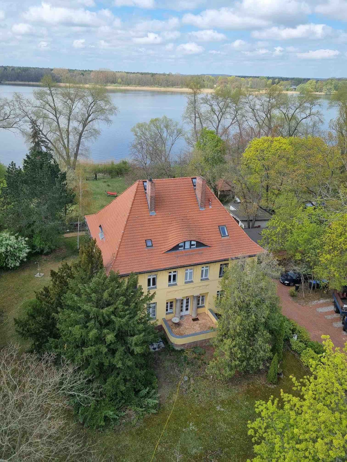 An aerial view displays a yellow villa with a distinctive red-tiled roof, surrounded by lush greenery. Nearby, the calm waters of a lake reflect the sky. A spacious garden is visible, providing a serene setting for relaxation.