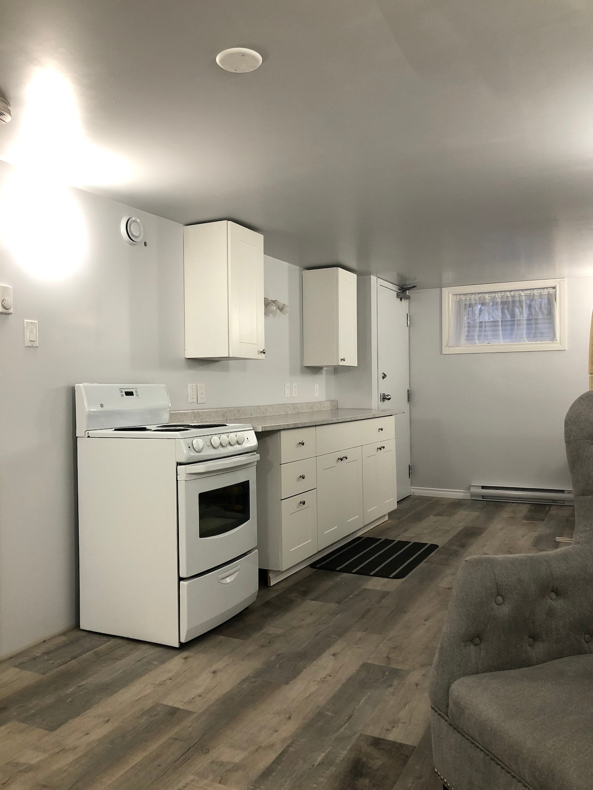 A compact kitchen area is seen, featuring a white stove and a countertop with storage cabinets above. A small window allows natural light to enter, illuminating the gray walls and laminate flooring. A rug lies on the floor near the stove, enhancing the functional space.