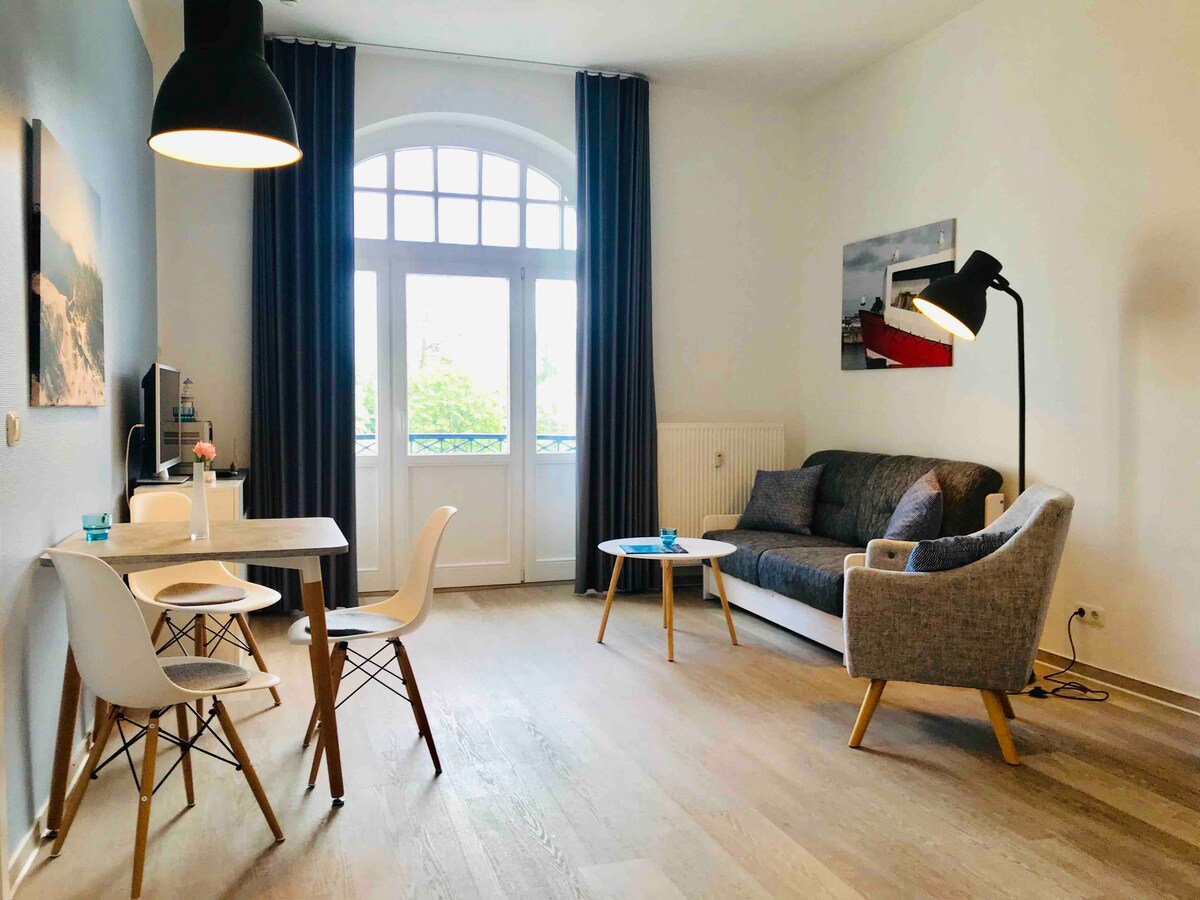 A bright living area features a comfortable sofa and modern chairs arranged around a small dining table. Tall windows allow natural light to fill the space, which is accented by stylish light fixtures and curtains, creating a relaxed atmosphere.