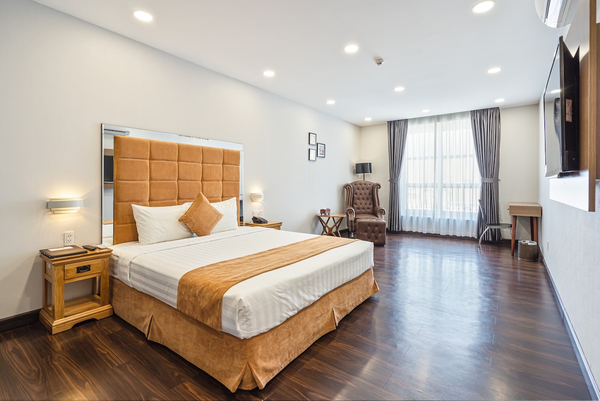 A spacious guest room features a comfortable bed with a beige headboard and a warm-toned bedspread. Natural light floods in through large windows, accentuated by gray curtains. A cozy seating area consists of a chair and a small table, with a mounted television on the wall.