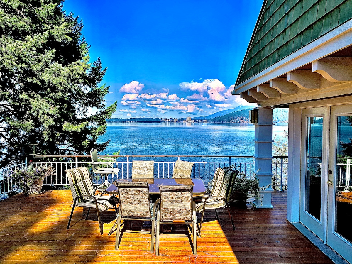 An expansive outdoor deck offers breathtaking lake views, furnished with a dining table surrounded by comfortable chairs. The natural wood flooring complements the lush greenery that frames the scene, creating a peaceful setting for al fresco meals or relaxation by the water.