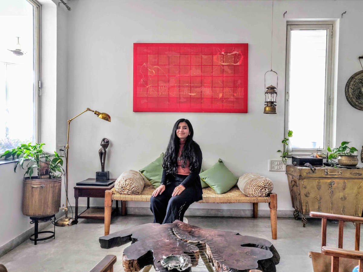 A cozy seating area features a light-colored bench adorned with green cushions. A large red artwork hangs on the wall above. Houseplants are positioned near the windows, allowing natural light to fill the space. Various decorative items are placed on a wooden table.