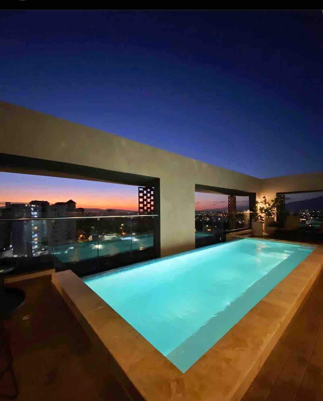 An infinity pool is positioned on a rooftop, reflecting the gradient of twilight colors. The surrounding areas feature modern architecture and a panoramic view of the city skyline. Soft lighting highlights the pool's edge, adding to the tranquil ambiance.