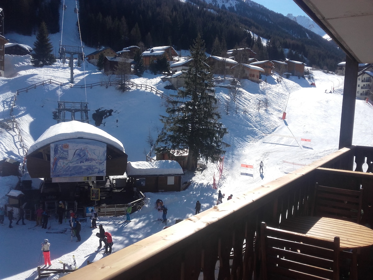 A view from a balcony overlooks a snowy landscape with skiers making their way down a slope. Nearby, a ski lift is visible, and rustic wooden chalets are scattered across the hillside, surrounded by evergreen trees under a clear blue sky.