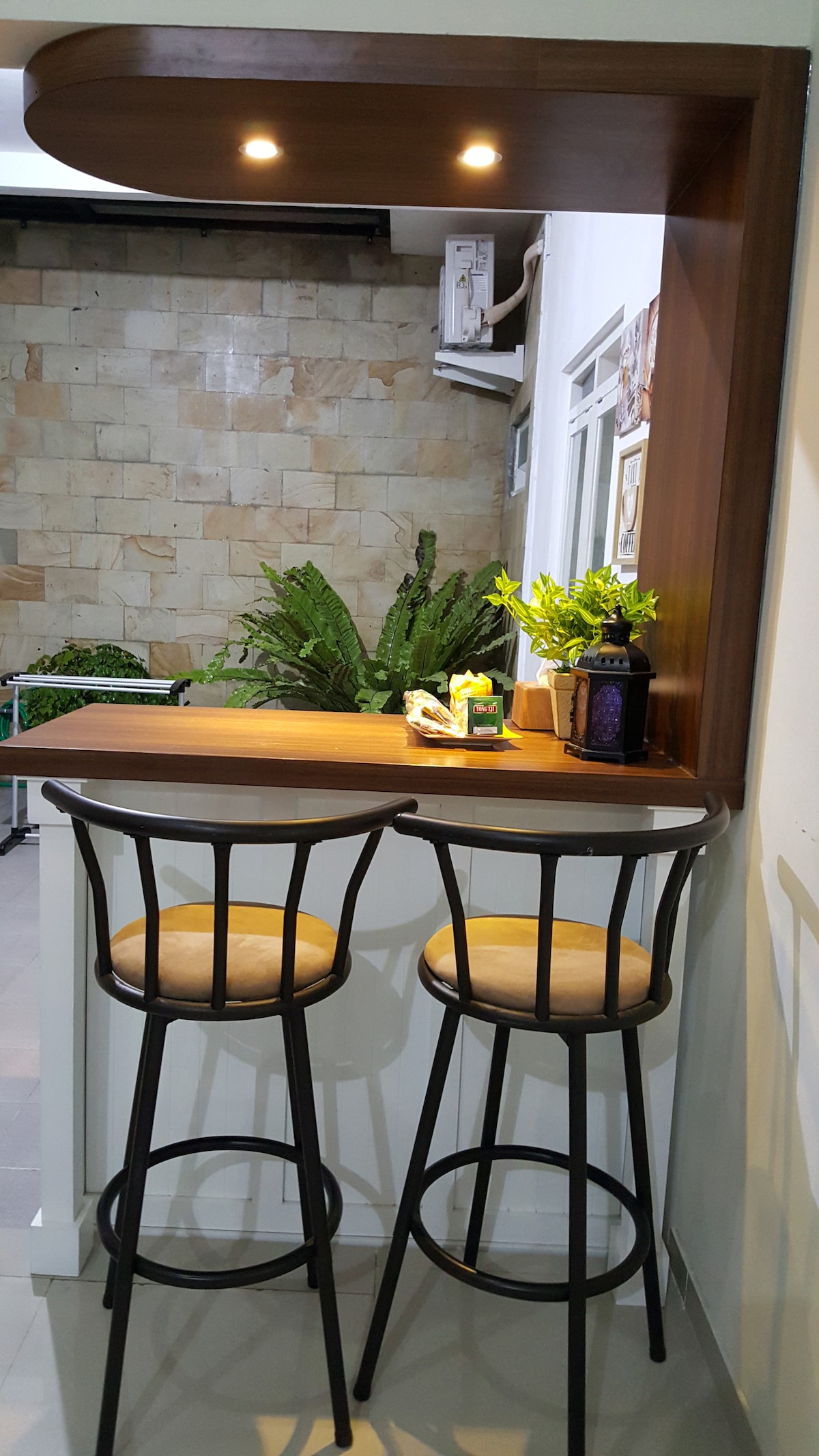 A minimalist bar area features a smooth wooden countertop with two black metal stools. Indoor plants are positioned on the countertop, while the backdrop consists of textured stone walls, adding a natural element to the space.