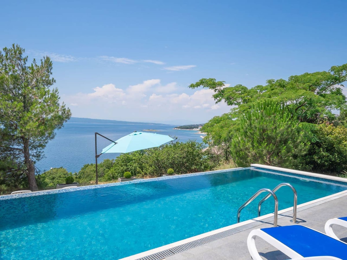 The image features a swimming pool with crystal-clear water, surrounded by greenery. An umbrella provides shade beside the pool, with a view of the sea and distant coastline visible under a clear sky.