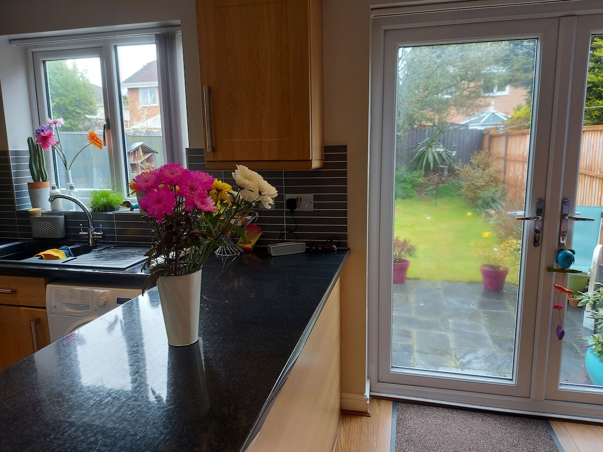 A modern kitchen features sleek dark countertops and light wood cabinetry. A vase of colorful flowers sits on the counter. Natural light enters through two windows and a glass door, which opens to a garden area with green grass and potted plants.