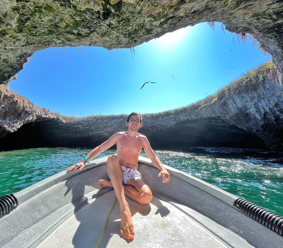 A sleek boat is positioned inside a natural rock formation, creating a striking arch. The vibrant blue sky and sunlight illuminate the scene, reflecting off the clear water. Lush greenery is visible on the rock edges, adding contrast to the surrounding natural beauty.