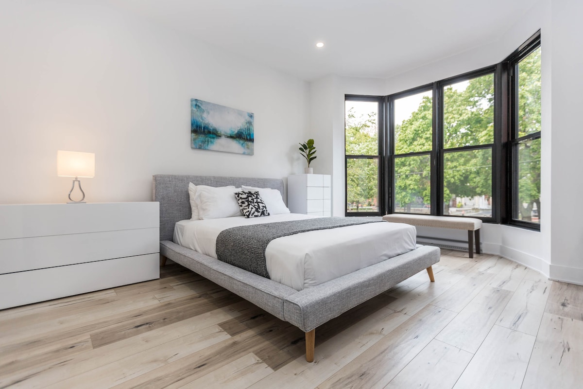 A spacious sleeping area is illuminated by natural light flowing through large bay windows. The light-colored wood floor complements the modern, minimalist furniture. A simple and elegant white dresser and a cozy bench are included, while a decorative piece of art hangs above the bed.