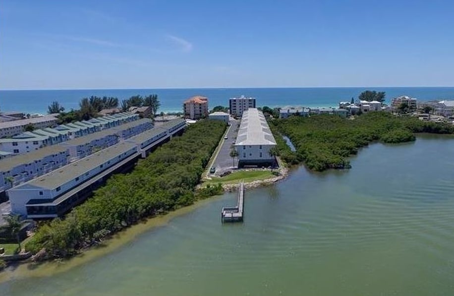 An aerial view captures the two-story complex surrounded by lush greenery, with the intracoastal waterway adjacent to the property. The coastline is visible in the distance, framing the serene waters and community structures that line the shore.