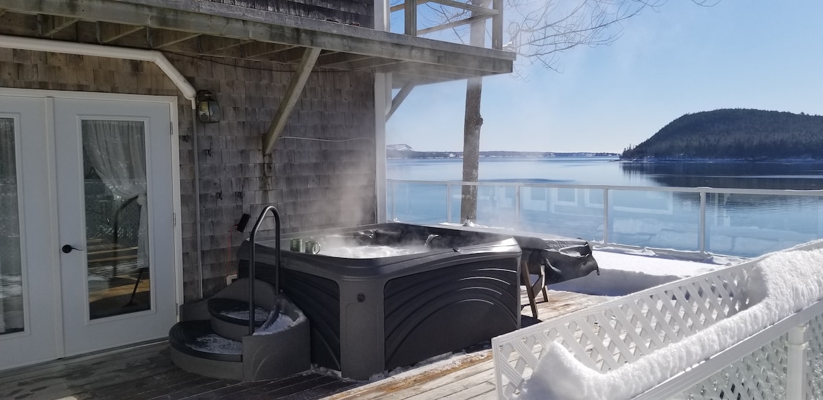 A hot tub with steam rising sits on a private deck, overlooking a calm water view that reflects the clear blue sky. A wooden staircase leads from the deck to a snowy landscape, enhancing the serene atmosphere.