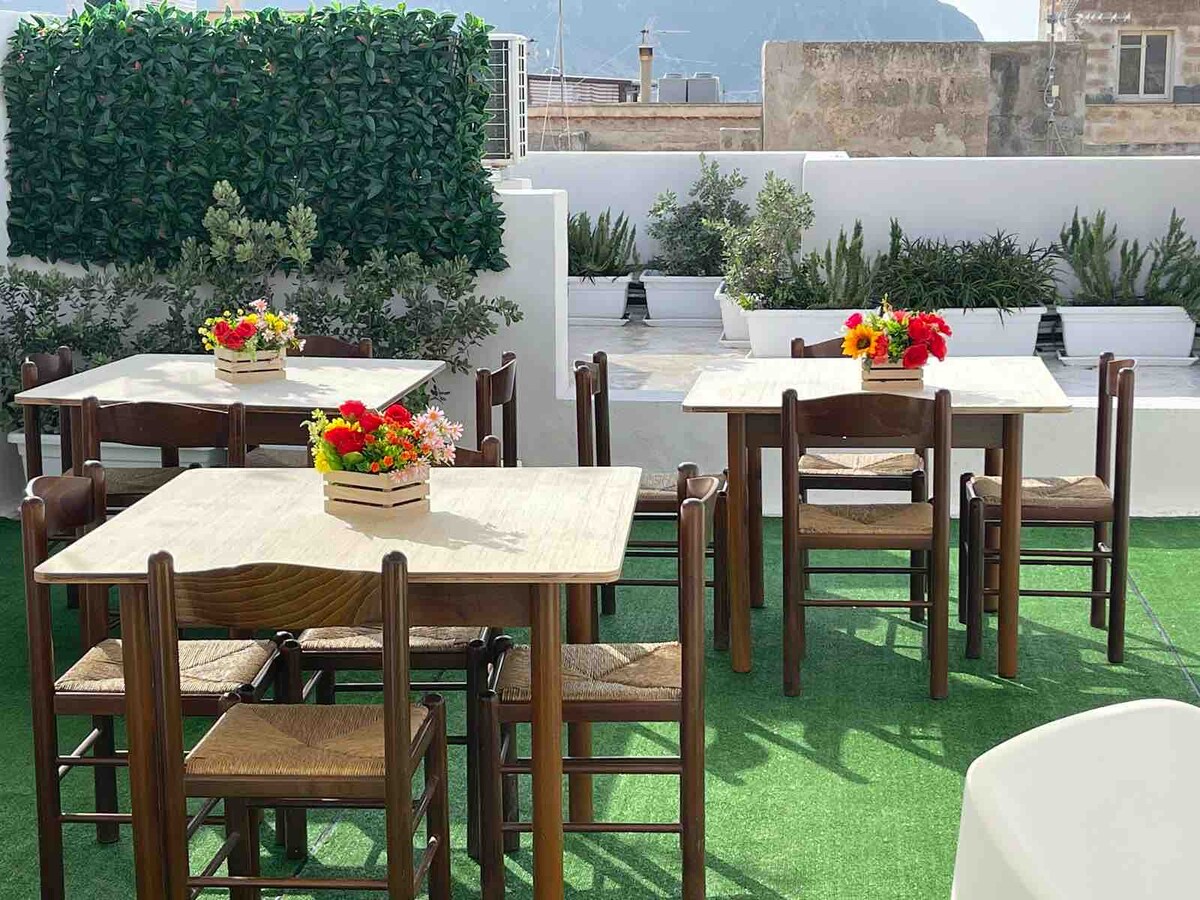 An outdoor terrace area displays several wooden tables accompanied by matching chairs. Each table is adorned with floral centerpieces, enhancing the green turf surrounding them. Landscaped plants and a low hedge can be seen in the background, contributing to a peaceful atmosphere.