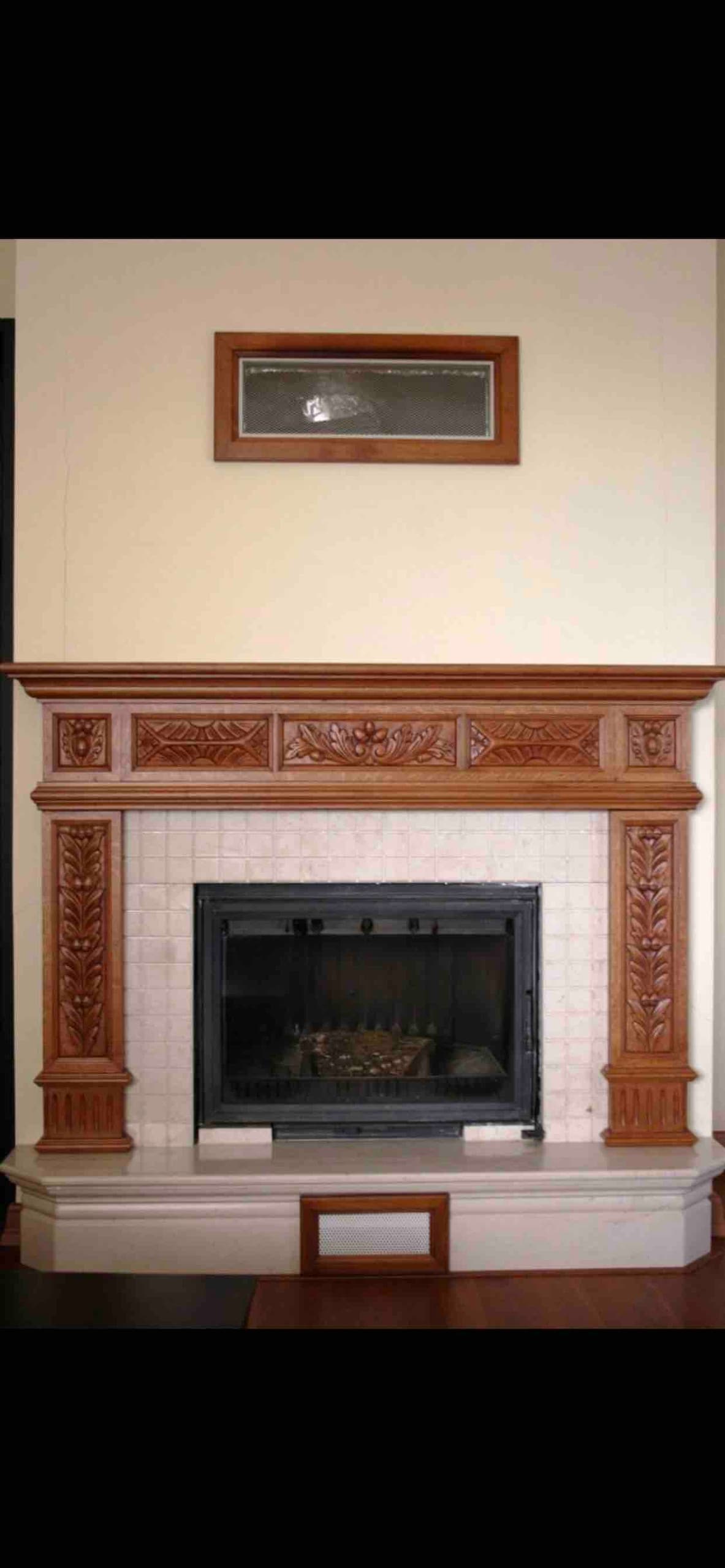 An intricately designed wooden fireplace serves as a focal point in the room. The mantel features detailed carvings, while a clean, lined hearth supports the fireplace opening. Above, a framed mirror adds depth to the space, contributing an elegant touch.