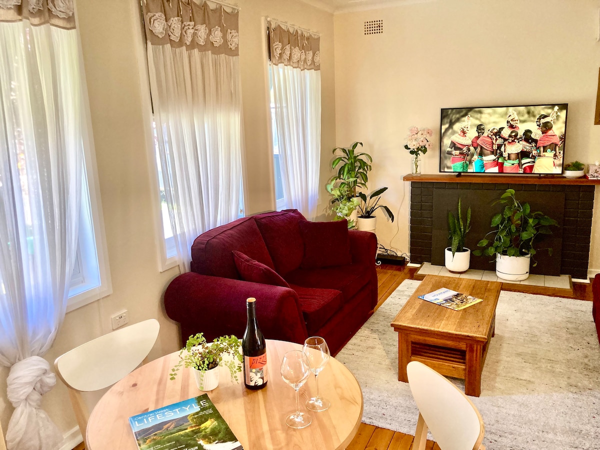 The lounge room features three long windows allowing natural light, with sheer curtains framing the view. A deep red sofa faces a coffee table, complemented by a small dining table with two chairs. A television mounted above a dark fireplace displays artwork, surrounded by potted plants.