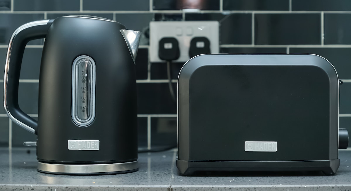 A black electric kettle and a black toaster are placed side by side on a kitchen countertop. The kettle features a clear water level indicator, while the toaster has a sleek design with a single slot for toasting. The background is accented with dark tiled backsplash.
