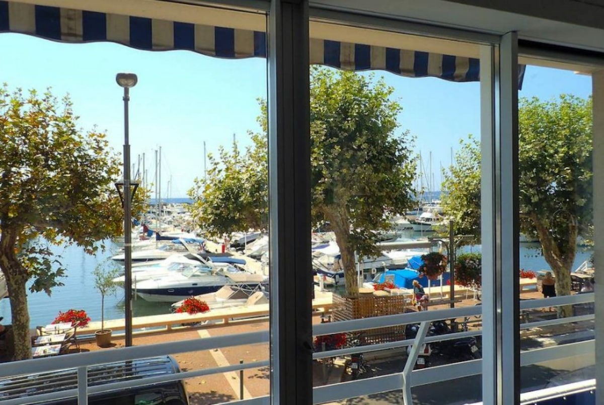 A view of the marina is visible through large glass windows, featuring various boats docked in the water. Trees with lush green foliage line the waterfront, creating a serene coastal atmosphere. The foreground includes a promenade with flower planters, enhancing the outdoor experience.
