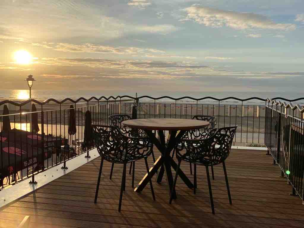 A large terrace features a circular wooden table surrounded by four intricately designed chairs. The sunset casts a warm glow over the sea, visible beyond a railing that complements the terrace's inviting atmosphere. Beach umbrellas and distant waves can also be observed.