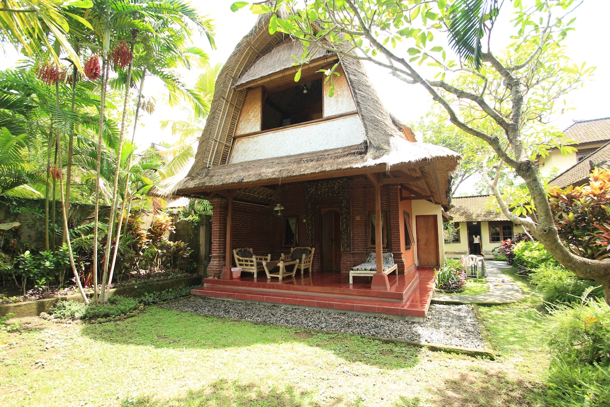 A charming two-story structure features a thatched roof and spacious verandah. Dining tables and chairs are positioned on the porch under natural shade from surrounding tropical plants. Lush gardens and a stone pathway enhance the serene outdoor setting.