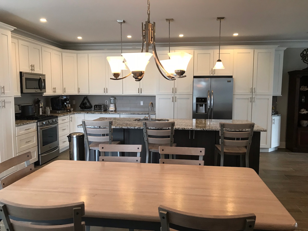 A modern kitchen features white cabinetry paired with granite countertops. Stainless steel appliances include a refrigerator and microwave. A dining table is situated in the foreground, surrounded by several wooden chairs. Multiple pendant lights illuminate the space above a central island.