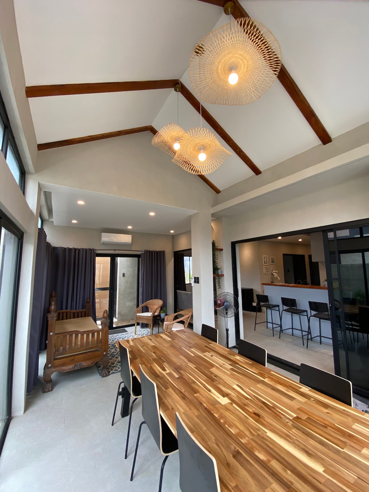 An open dining area is highlighted by a long wooden table surrounded by sleek black chairs. The high ceiling features unique woven light fixtures, while large glass doors offer views of the outside. Soft curtains frame the windows, creating a bright and airy environment.