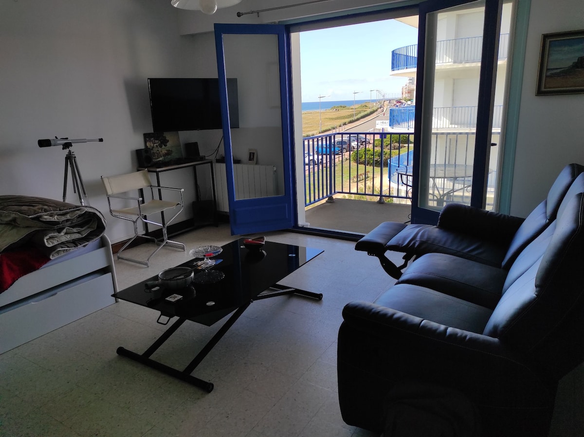 A living area features a plush black sofa and a sleek glass coffee table, complemented by a television mounted on the wall. Large sliding glass doors open to a balcony with views of the ocean, allowing natural light to fill the space.