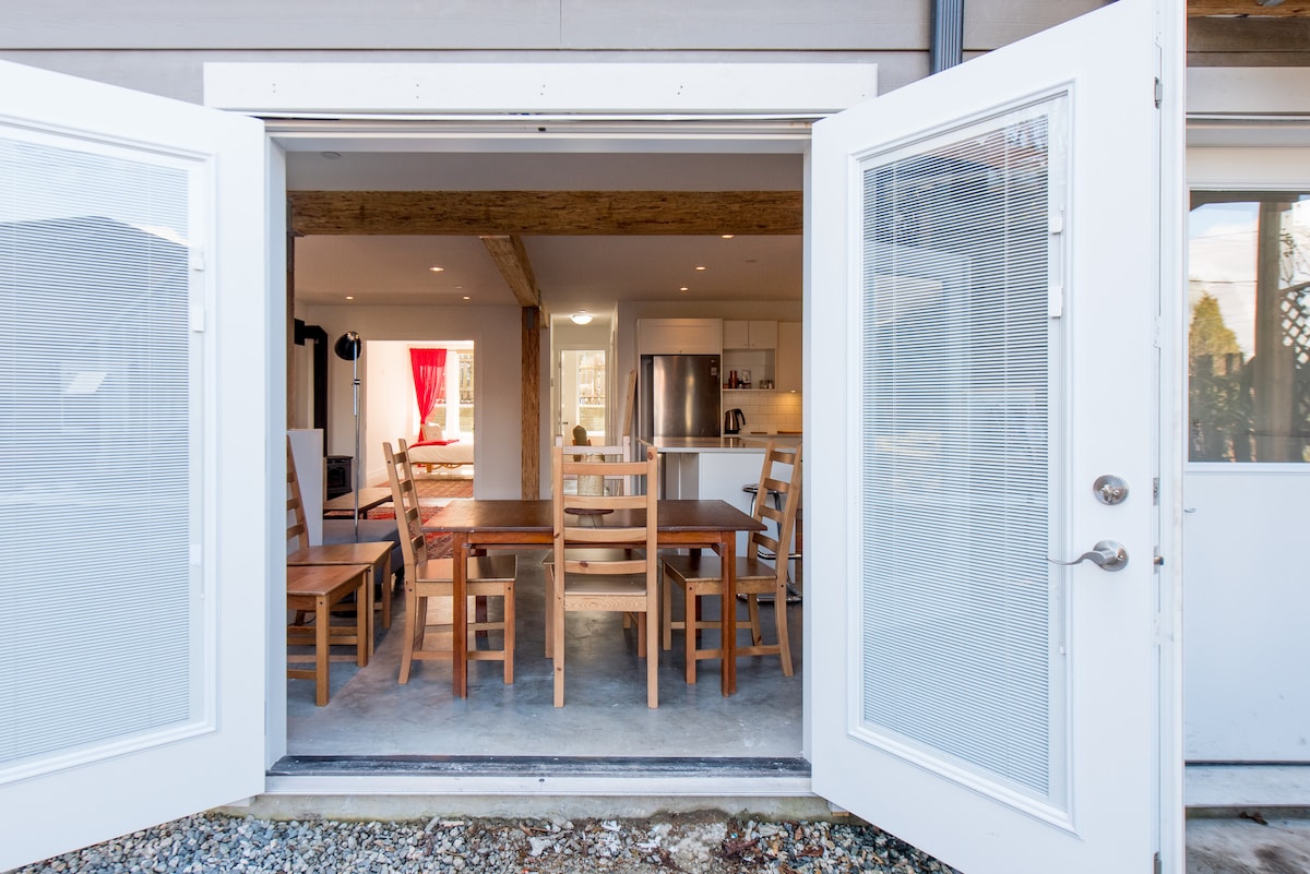 The image shows an open door leading to a bright indoor dining area with a large table surrounded by multiple wooden chairs. Light wood beams are visible above, enhancing the spacious feeling, while the kitchen area can be seen in the background.