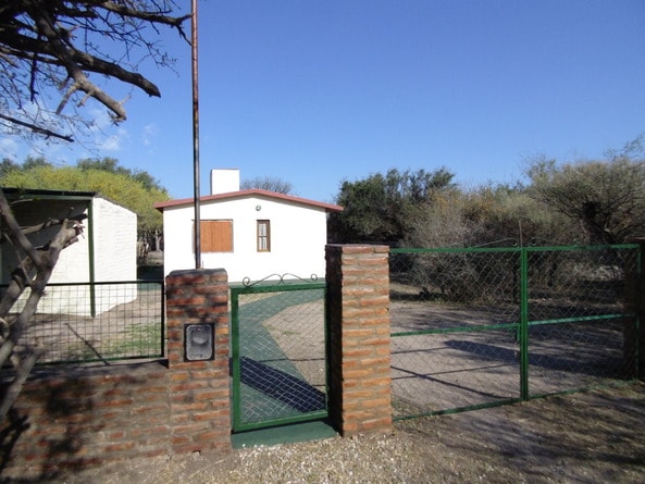 A white house is visible behind a green gate, surrounded by native trees and shrubs. A brick fence encloses the property, providing a secure entrance. The clear blue sky above complements the tranquil setting.
