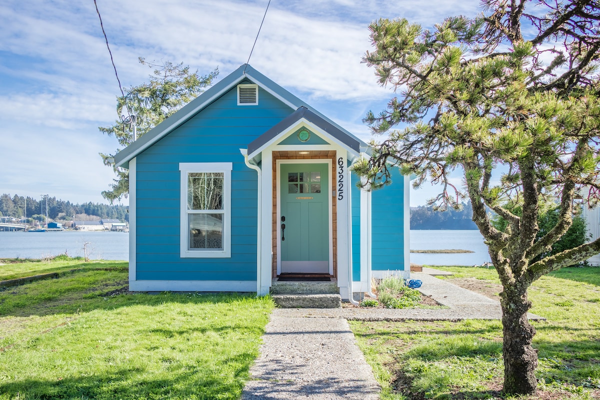 A cozy blue cottage is presented with a welcoming front porch and a light pathway leading to the entrance. Large windows allow natural light to brighten the interior. The lush green lawn contrasts with the vibrant exterior, while the water and trees create a serene backdrop.